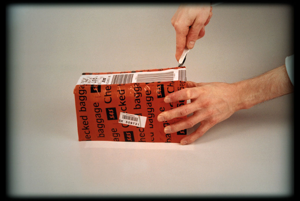 The Trip To America, film still, paper box covered in tape labelled Checked Bagage is being opened by a knife.