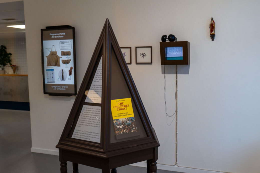 Museum vitrine with a copy of The Childfree Christ, a book by Théophile de Giraud. Video screens in the background. Art installation by Karl & Carl.