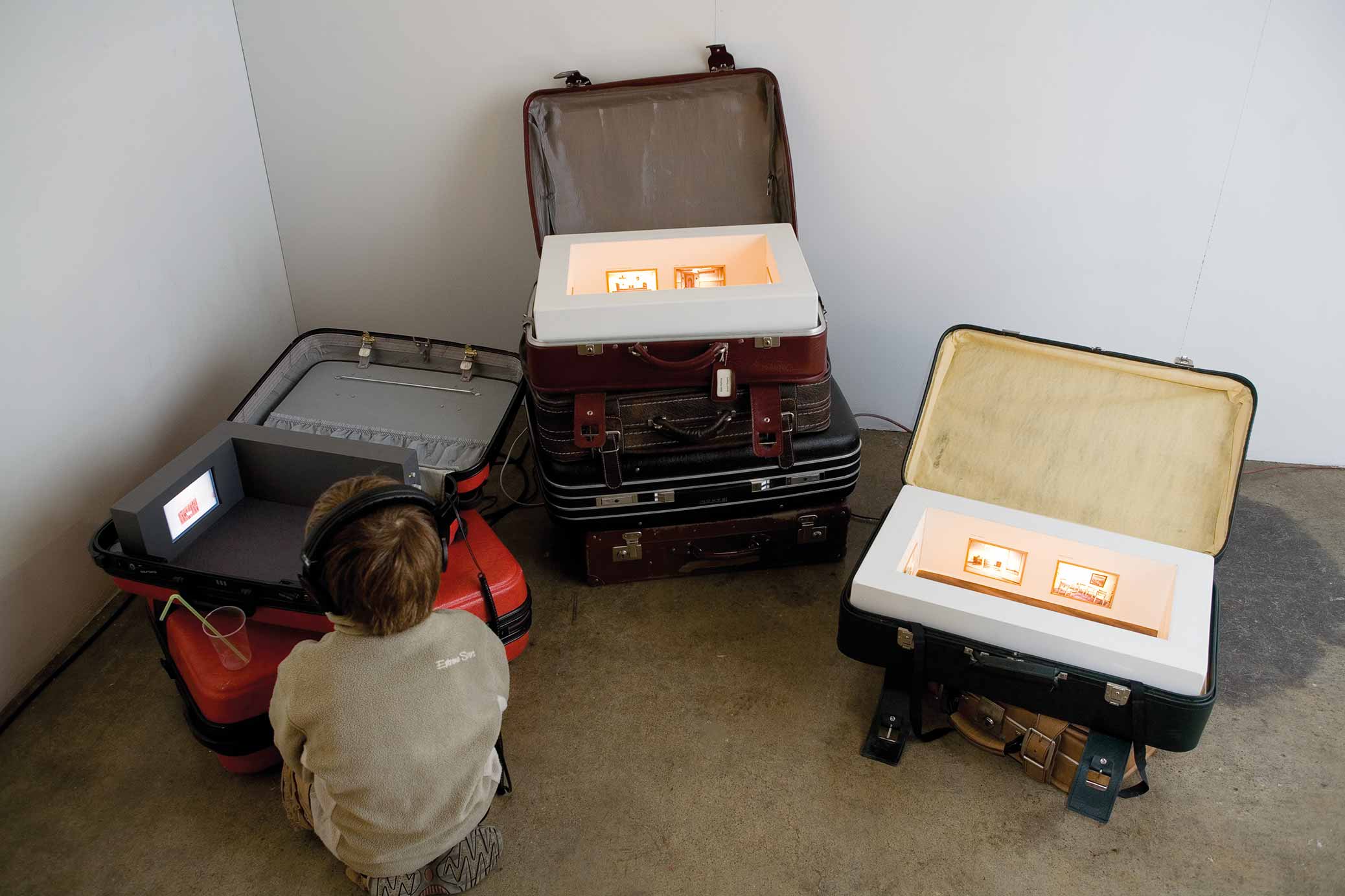 Resväsksetyder, installation med miniatyrutställningar inuti resväskor. Pojke tittar på video.